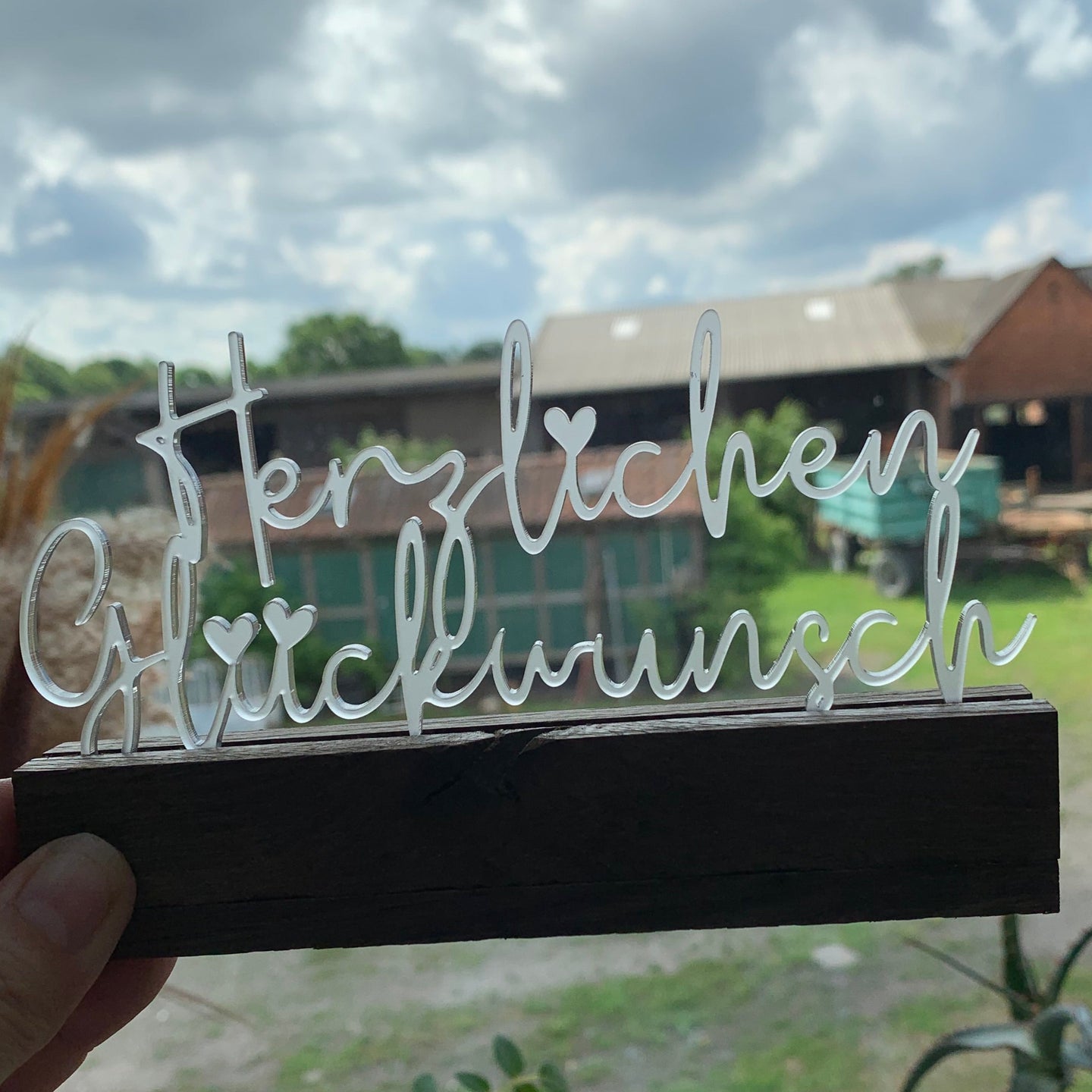 Ein handgehaltener Acryl Aufsteller von Easy-Sticks mit dem Schriftzug 'Herzlichen Glückwunsch', vor einem hübschen Hintergrund im Freien. Ideal, um jedem Anlass eine besondere Note zu verleihen.