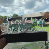 Ein handgehaltener Acryl Aufsteller von Easy-Sticks mit dem Schriftzug 'Herzlichen Glückwunsch', vor einem hübschen Hintergrund im Freien. Ideal, um jedem Anlass eine besondere Note zu verleihen.