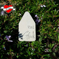 Ein graues Namensschild mit dem Wort "Viel Glück", umgeben von grünen Pflanzen und lila Blüten, ein weiteres kreatives Projekt aus der Plotterdatei "Kerzensprüche für jeden Anlass 2" von easy-sticks.
