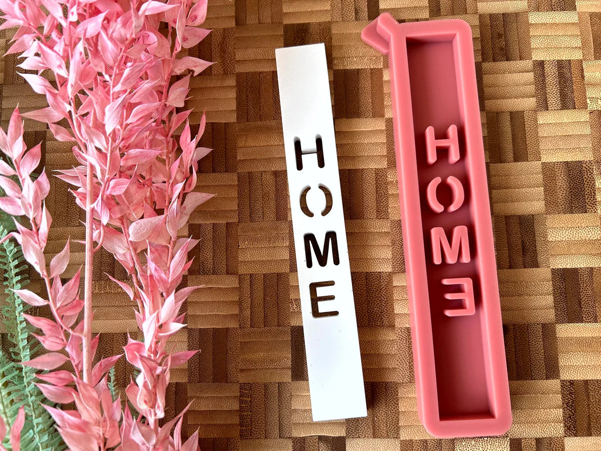 Bunte Silikonform "HOME Schriftzug" von easy-sticks, präsentiert neben einer eleganten weißen Form auf einer Holzunterlage, ideal für kreative DIY-Projekte in der He dekor.