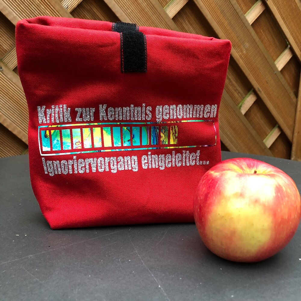 Eine rote Tasche mit dem Aufdruck 'Kritik zur Kenntnis genommen ... Ignoriervorgang eingeleitet' von easy-sticks, zusammen mit einem frischen Apfel, ideal für kreative und farbenfrohe Designs.