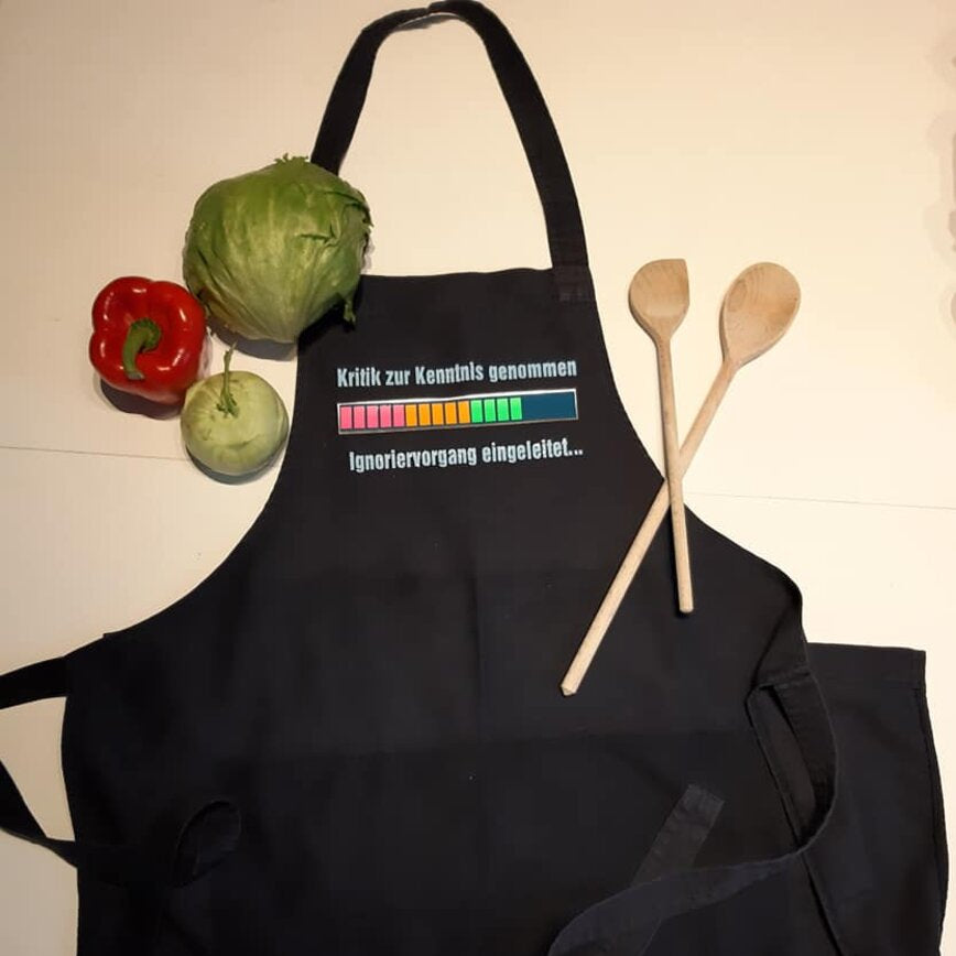 Eine dunkle Kochschürze mit dem Aufdruck 'Kritik zur Kenntnis genommen ... Ignoriervorgang eingeleitet' von easy-sticks, umgeben von frischen Zutaten, darunter Gemüse und Kochlöffel.