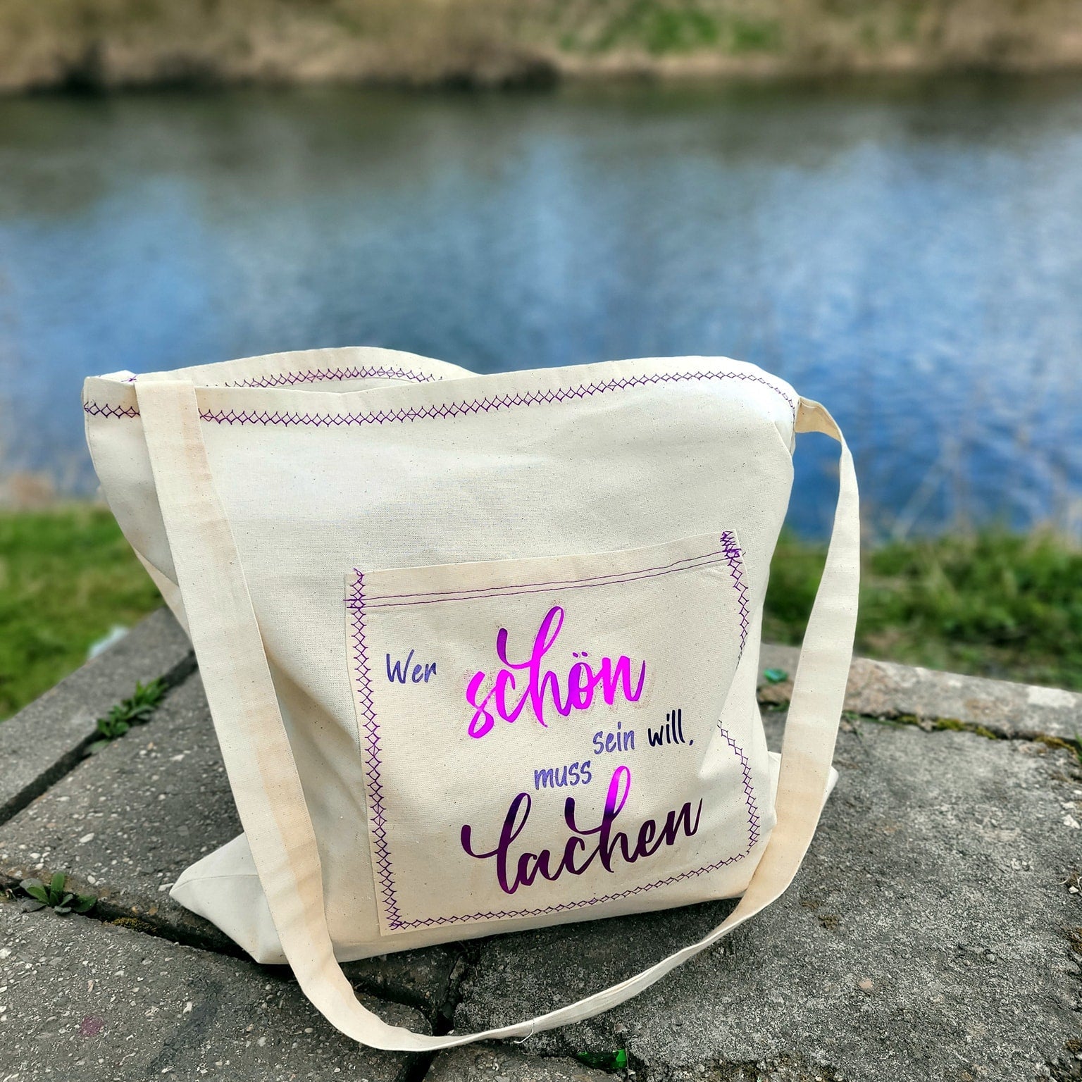 Stilvolle Tragetasche mit dem Design "Wer schön sein will muss lachen" von easy-sticks, aufgenommen am Wasser für kreative Nutzung.
