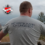 Mann mit grauem T-Shirt, das das Design "Mit Kritik kann ich umgehen - und mit einer Axt" trägt. Perfekt für persönliche DIY-Ideen mit der Plotterdatei von easy-sticks.