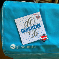 Eine blaue Tasche mit einem Geschenk, das eine Karte mit der Aufschrift 'GESCHENK' trägt. Perfekt für kreative Bastelideen, inspiriert von der Monogram 'G' Plotterdatei von easy-sticks.