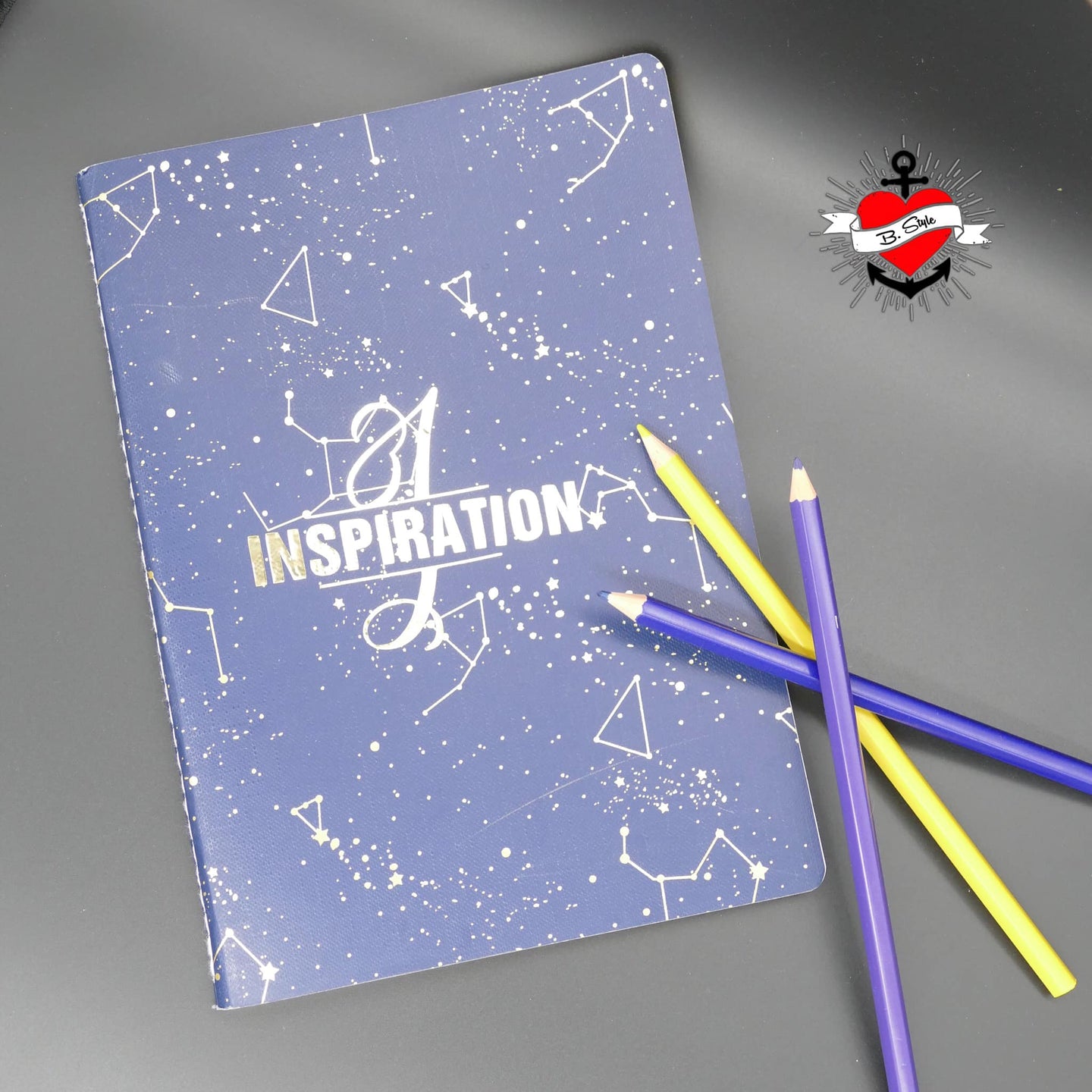 Notizbuch mit dem Titel INSPIRATION und dem Monogramm "I", gestaltet von easy-sticks, umgeben von bunten Buntstiften auf einer grauen Oberfläche.