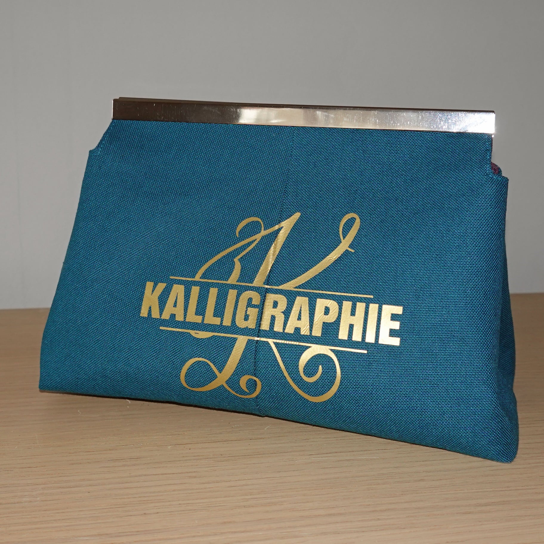 Monogramm 'K' Plotterdatei von easy-sticks gestaltet auf einer türkisfarbenen Tasche mit goldenem Schriftzug 'Kalligraphie'. Ideal für persönliche Accessoires.