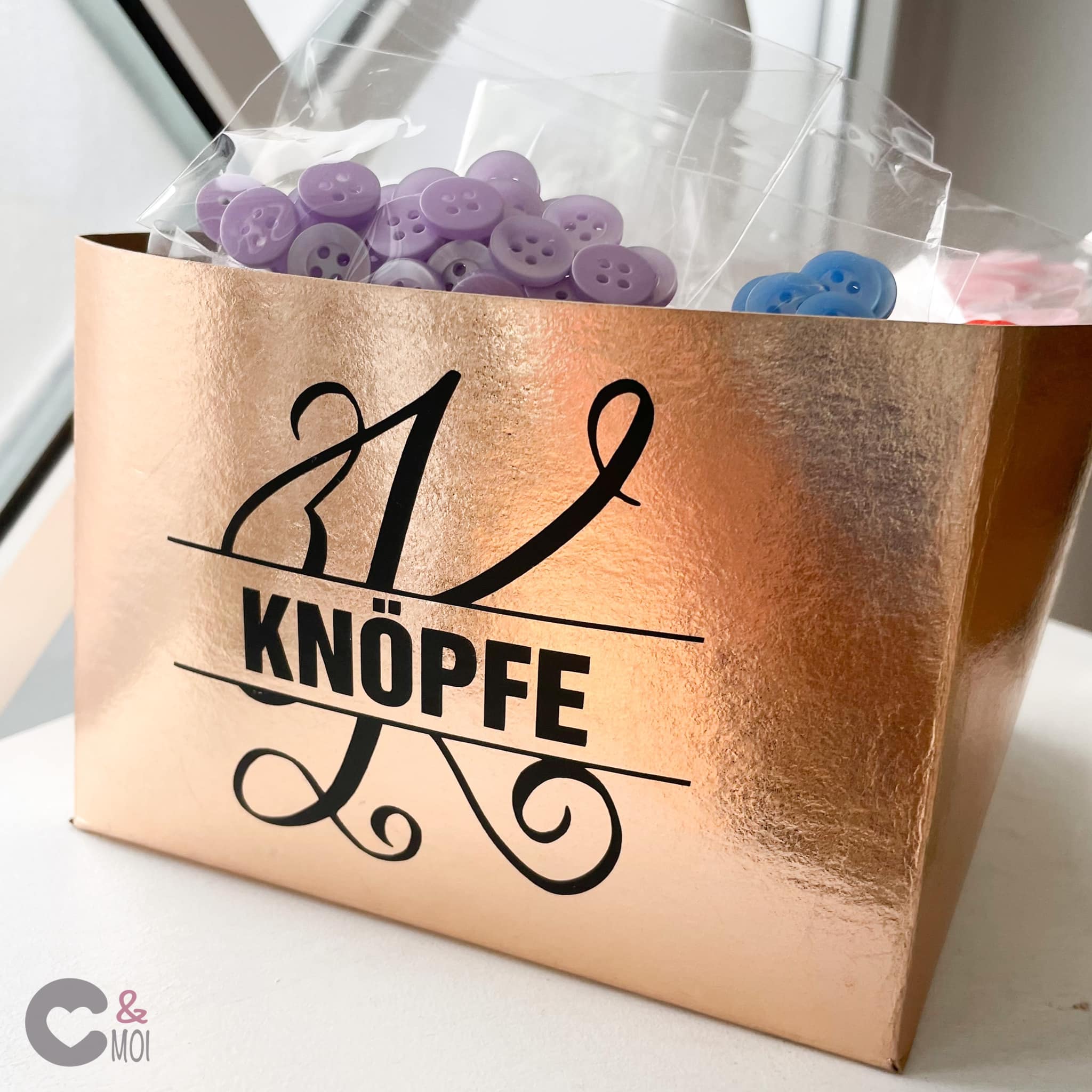 Monogramm 'K' Plotterdatei von easy-sticks auf einer glänzenden roségoldenen Box mit lila Knöpfen. Perfekt für kreative DIY-Projekte.