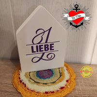 Dekorativer Holzblock mit der Monogramm "L" Plotterdatei von easy-sticks, der die Worte "1 LIEBE" in leuchtendem Lila zeigt, platziert auf einem bunten Untersetzer.