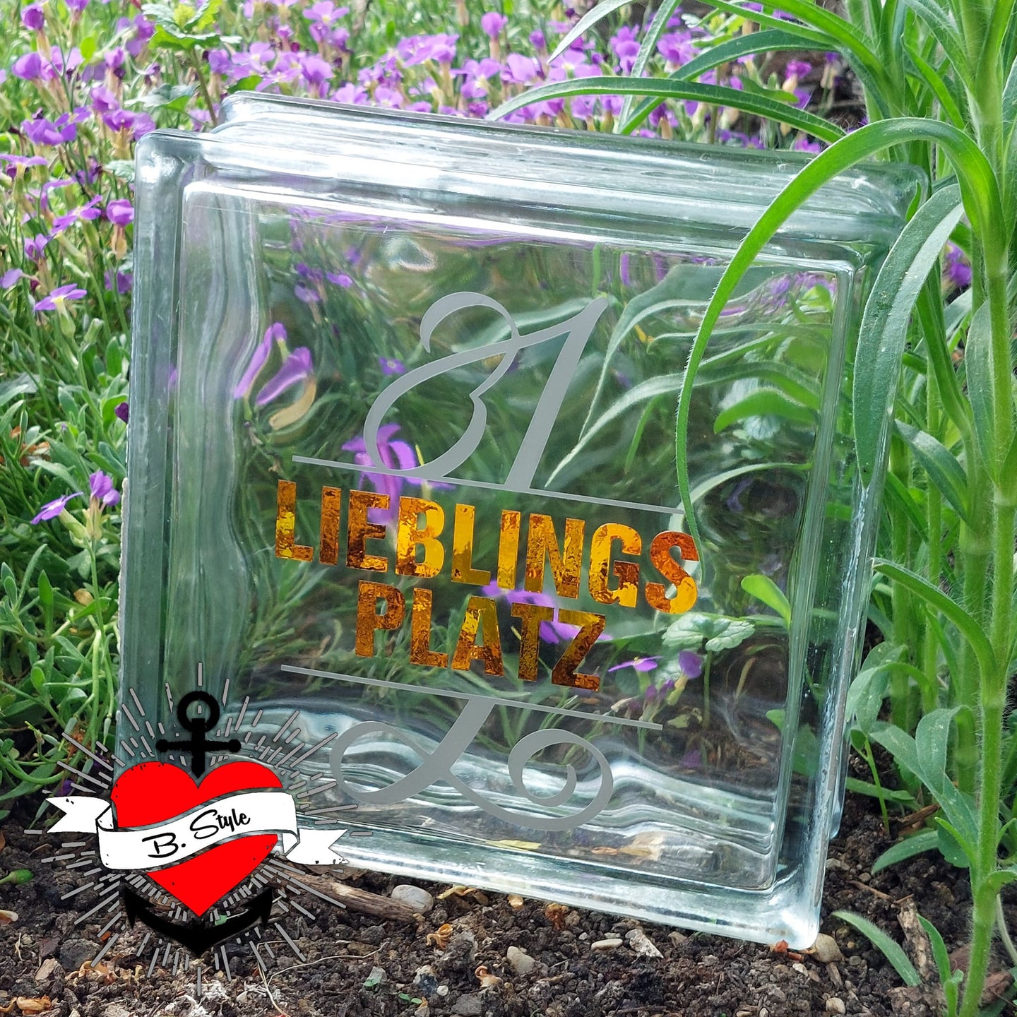 Glasblock mit der Monogramm "L" Gestaltung von easy-sticks, auf dem die Schriftzüge "1 LIEBLINGS PLATZ" in glänzenden Farben zu sehen sind und von bunten Blumen umgeben ist.