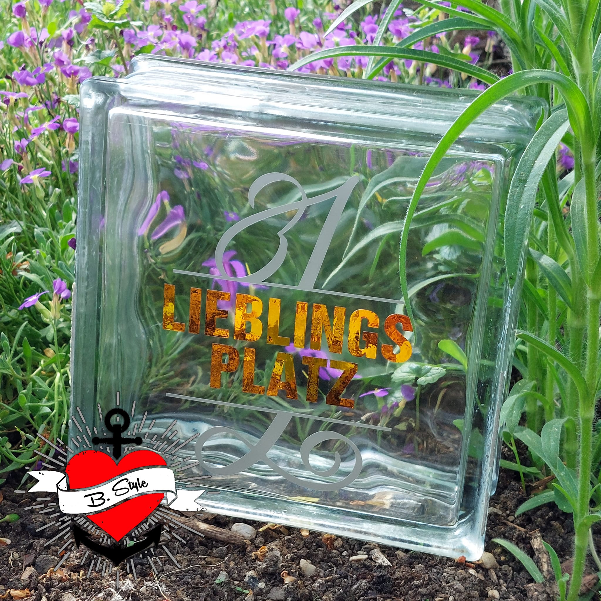 Glasblock mit der Monogramm "L" Gestaltung von easy-sticks, auf dem die Schriftzüge "1 LIEBLINGS PLATZ" in glänzenden Farben zu sehen sind und von bunten Blumen umgeben ist.