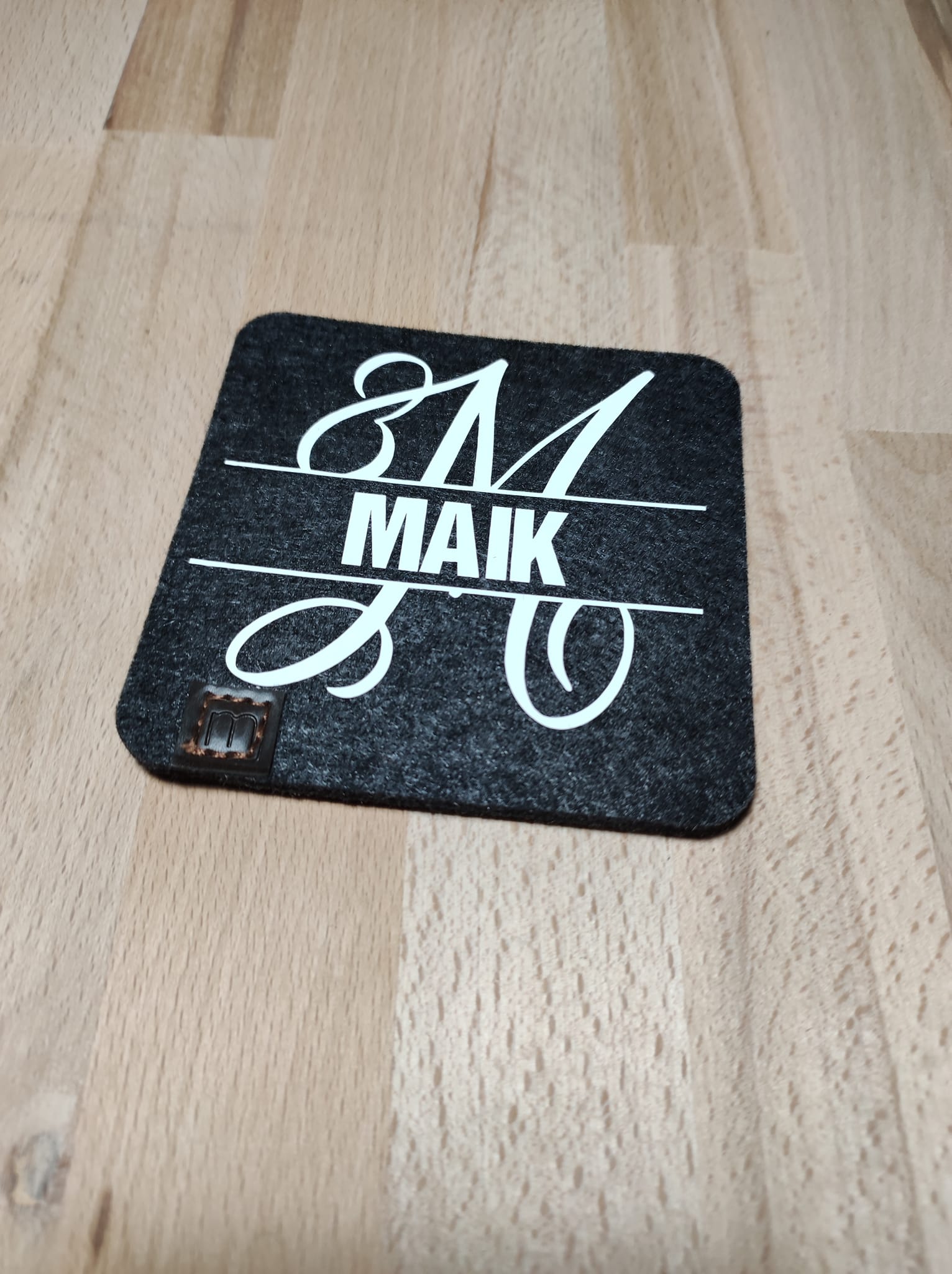 Monogramm "M" Plotterdatei auf einem schwarzen Filzuntersetzer mit dem Namen "MAIK", designt von easy-sticks. Perfekte Wahl für DIY-Projekte.