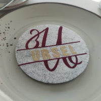 Stilvoller Tischuntersetzer mit einem personalisierten Monogramm "U" von easy-sticks, dekoriert mit dem Namen "URSEL" in glitzernden goldenen Buchstaben auf grauem Filz, eine kreative Idee für besondere Anlässe.