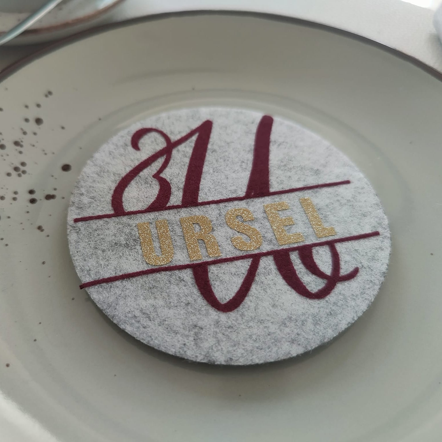 Stilvoller Tischuntersetzer mit einem personalisierten Monogramm "U" von easy-sticks, dekoriert mit dem Namen "URSEL" in glitzernden goldenen Buchstaben auf grauem Filz, eine kreative Idee für besondere Anlässe.