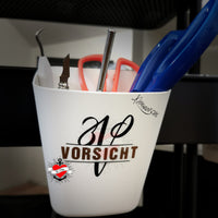Monogramm 'V' Plotterdatei von easy-sticks, auf einem weißen Behälter mit Schreibwerkzeugen wie Scheren, Klingen und einem Stift, gekennzeichnet mit dem Hinweis 'VORSICHT'.