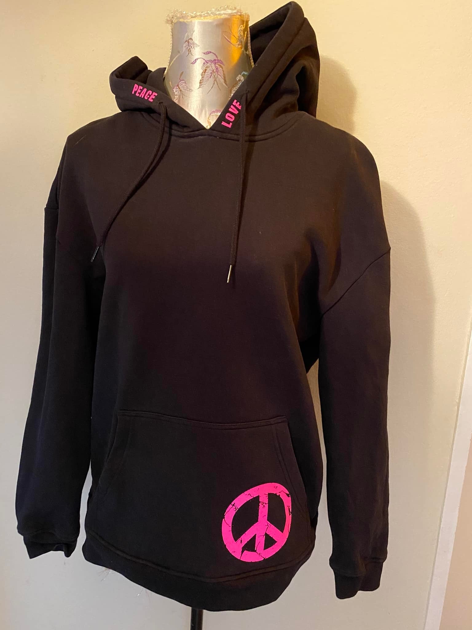 Eine schwarze Kapuzenshirt mit pinken Schriftzügen "PEACE" und "LOVE" sowie einem Peace-Symbol, designt aus der "PEACE" Grunge Schriftart Plotterdatei von easy-sticks. Perfekt für trendy Outfits! Plotterdatei.