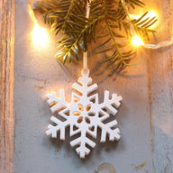 Schneeflocke zum Aufhängen von easy-sticks, dekorative weiße Schneeflocke aus Silikon, umgeben von Tannenzweigen und warmem Licht, ideal für festliche Dekoration und Geschenkverzierungen.