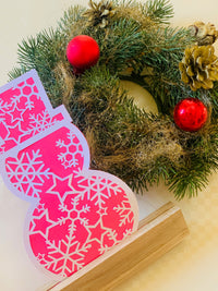 Schneemann Plotterdatei von easy-sticks, dargestellt in leuchtendem Pink, platziert vor einem festlichen Weihnachtskranz mit roten Dekorationen. Ideal für kreative Winterprojekte.