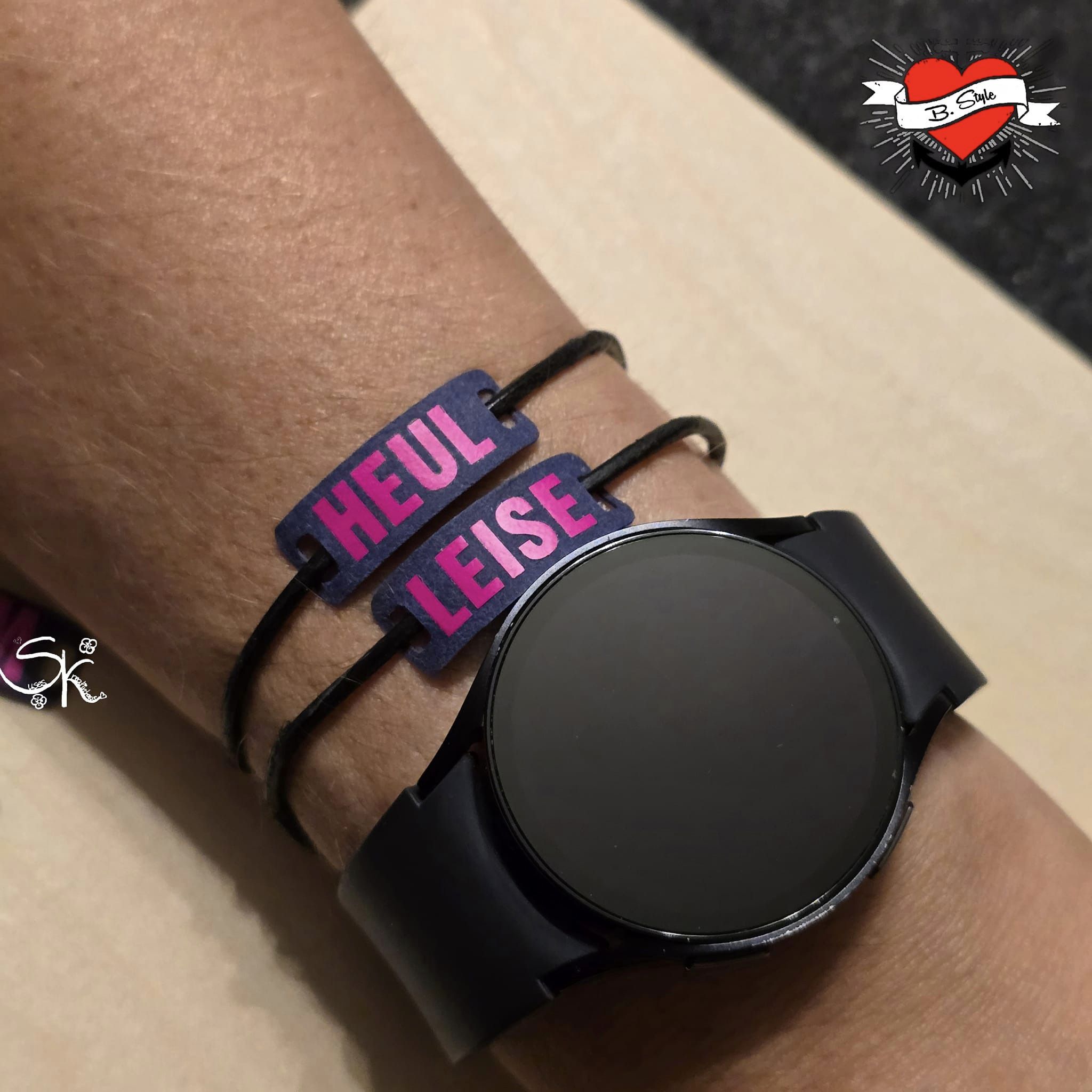 Armband mit personalisierten Schildern "HEUL" und "LEISE" auf einem Handgelenk, zusammen mit einer Smartwatch. Entdecke die "Armband und Schnürsenkel Schilder mit Wörtern" Plotter und Laser Datei von easy-sticks für kreative Projekte.