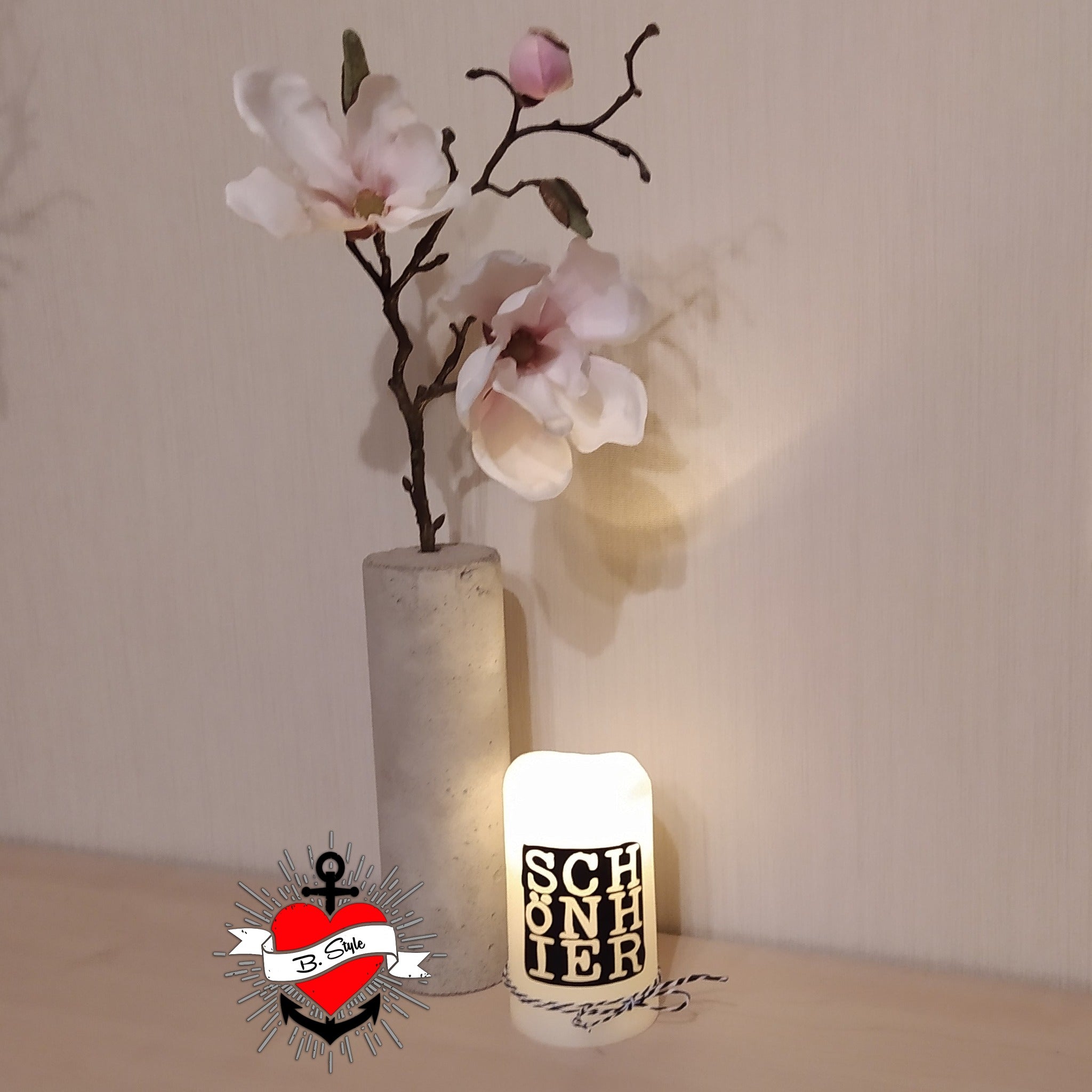 Dekoarrangement mit einer eleganten, künstlichen Magnolienblüte und einer leuchtenden Kerze im Hintergrund, die das Design der "SCHÖN HIER" Plotterdatei von easy-sticks optimal in Szene setzt und kreative Bastelideen inspiriert.