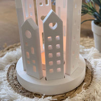 Kreative Windlichtgestaltung mit Easy-Sticks Silikon Grundform für Windlicht - Rund. Das Bild zeigt ein weißes Windlicht mit detaillierten Hausformen und einem sanften Lichtschein, ideal für gemütliche Dekoration.