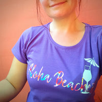 Frau mit lila T-Shirt, das den Aufdruck "Aloha Beaches" zeigt, vor einem rosa Hintergrund. Perfekt für den Sommerurlaub. Entdecke die "Sommer und Urlaub Sprüche" Plotterdatei von easy-sticks für kreative Projekte.