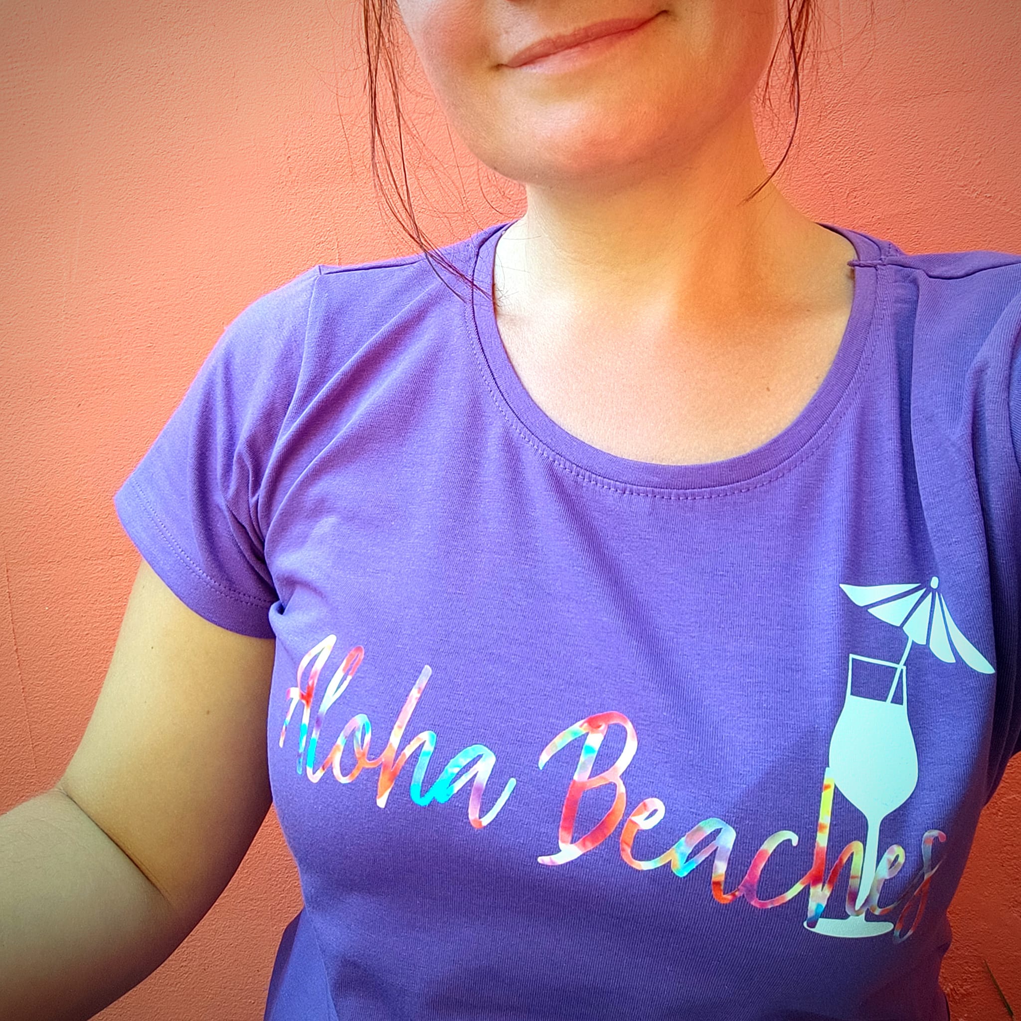 Frau mit lila T-Shirt, das den Aufdruck "Aloha Beaches" zeigt, vor einem rosa Hintergrund. Perfekt für den Sommerurlaub. Entdecke die "Sommer und Urlaub Sprüche" Plotterdatei von easy-sticks für kreative Projekte.