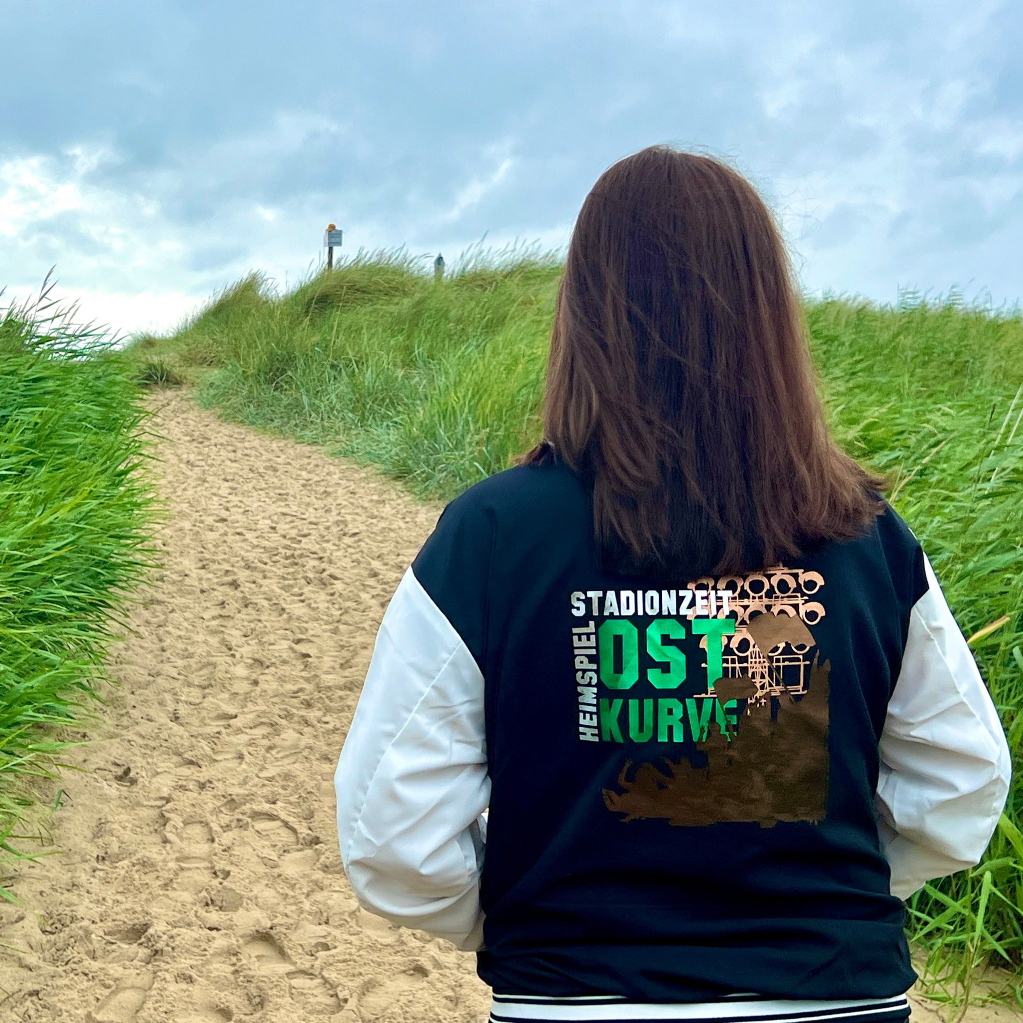 Frau mit Stadionzeit Jacke von easy-sticks auf einem Sandweg, ideal für Fußballfans, zeigt Stolz und Leidenschaft für die Stadionzeit.