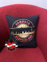 Stempel Skyline Set 16 Landeshauptstädte von easy-sticks, ein dekoratives Kissen mit dem Skyline-Motiv von Hamburg auf einem schwarzen Stoff vor einem roten Sessel.