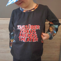 Ein Kind trägt ein T-Shirt mit dem Aufdruck "TAGSÜBER ZIRKUS ABENDS THEATER" von easy-sticks, eine bezaubernde Plotterdatei für kreative Mode.