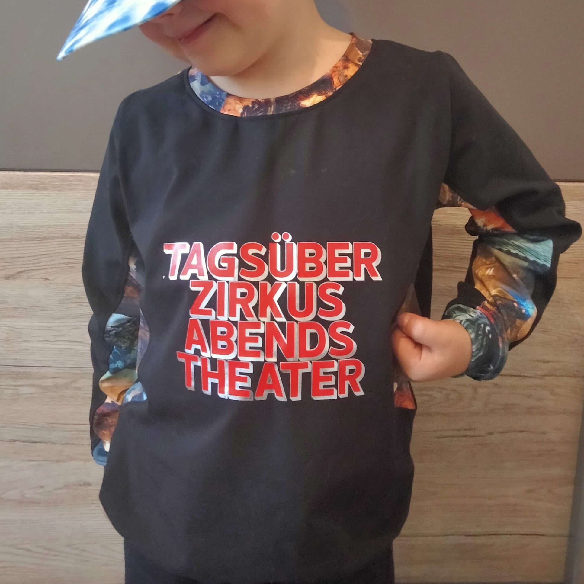 Ein Kind trägt ein T-Shirt mit dem Aufdruck "TAGSÜBER ZIRKUS ABENDS THEATER" von easy-sticks, eine bezaubernde Plotterdatei für kreative Mode.