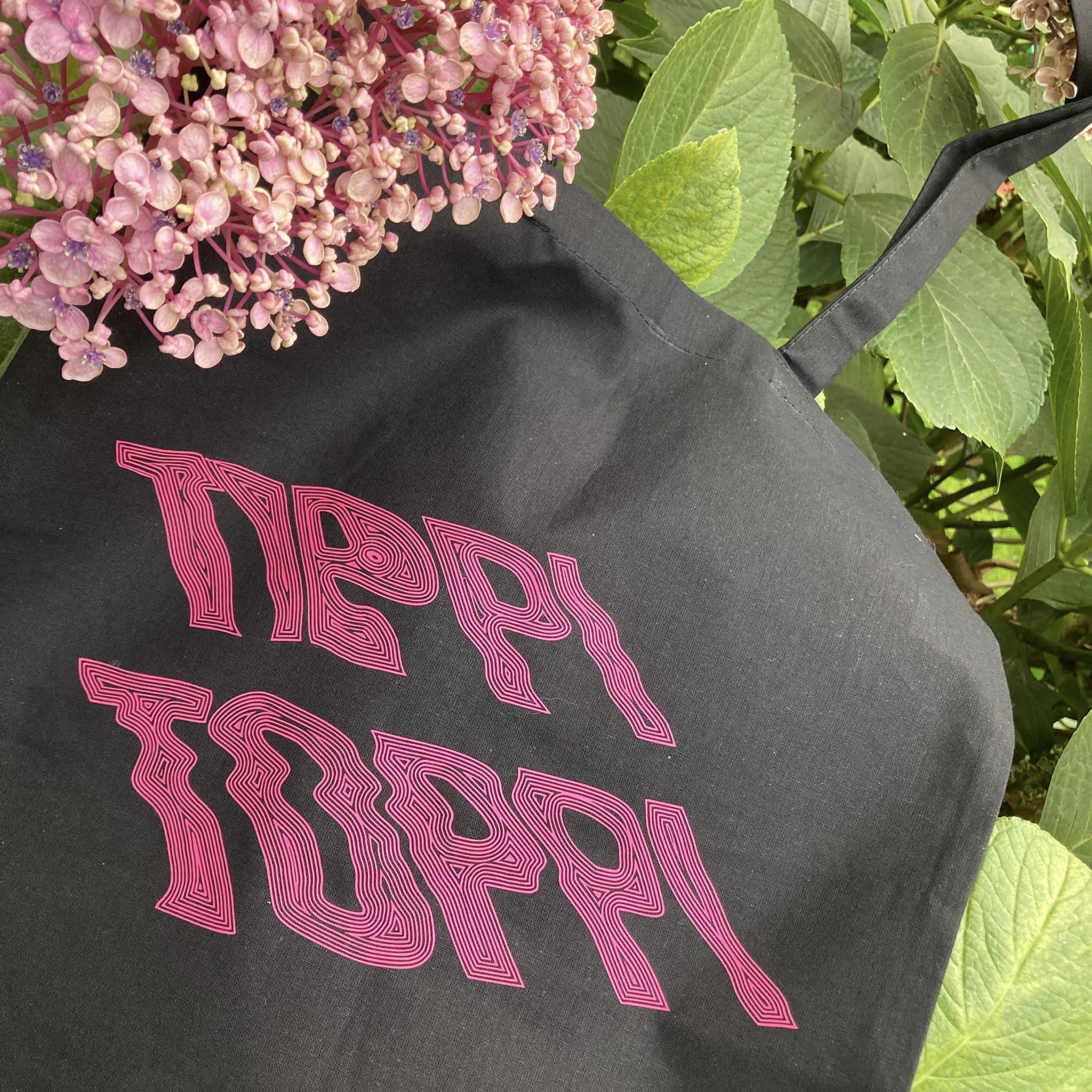 Schwarze Tragetasche mit dem pinken Schriftzug "TIPPI TOPPI" von easy-sticks, umgeben von blühenden Pflanzen.