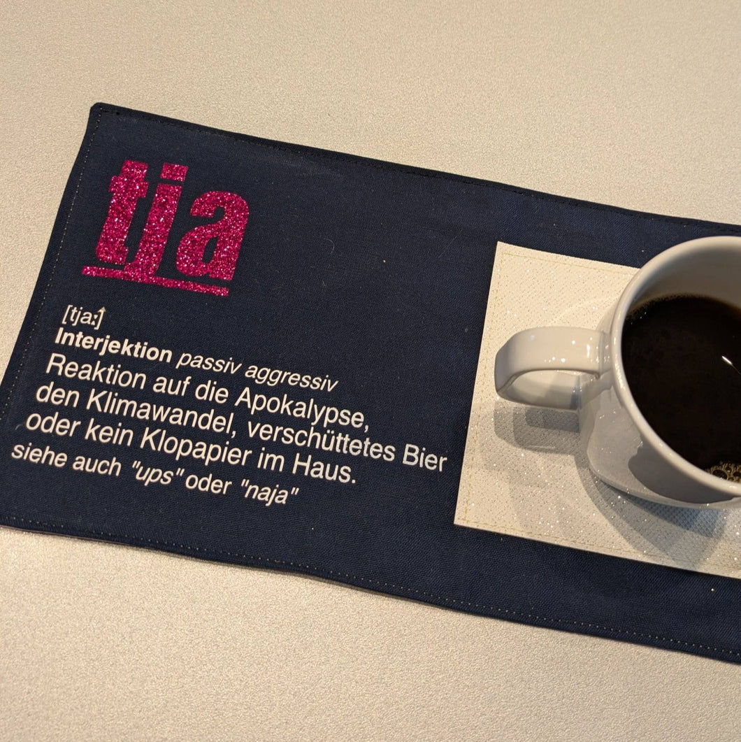 Kreatives Tischset mit dem Wort 'tja' in glitzerndem Pink, das auf einer dunklen Unterlage gedruckt ist, inklusive einer Tasse Kaffee, angeboten von easy-sticks als Teil der 'Tja' Plotterdatei.