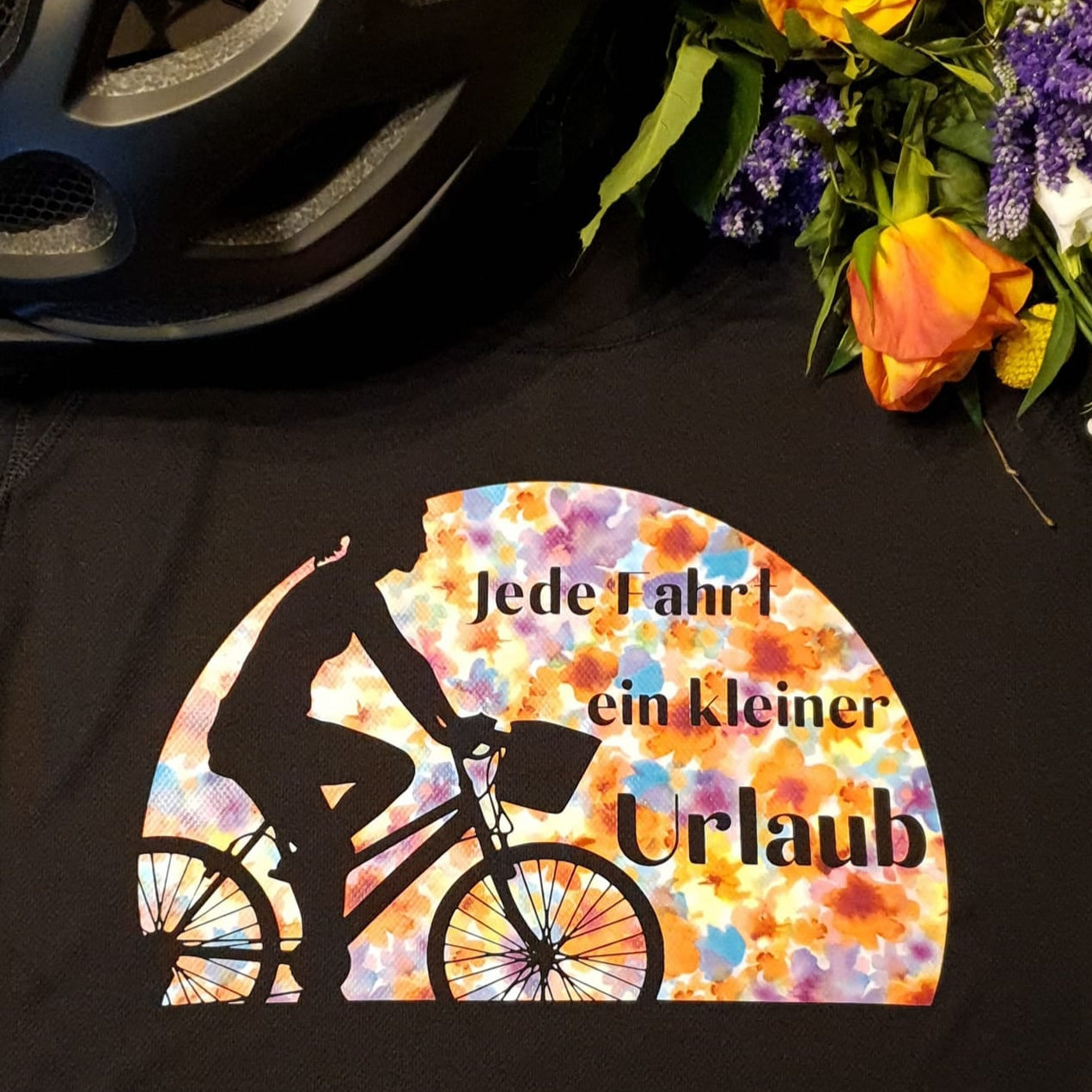 Schwarzes Shirt mit dem fröhlichen Aufdruck 'Jede Fahrt ein kleiner Urlaub' von easy-sticks, umgeben von einem Fahrradhelm und frischen Blumen, ideal für Radfahrer.