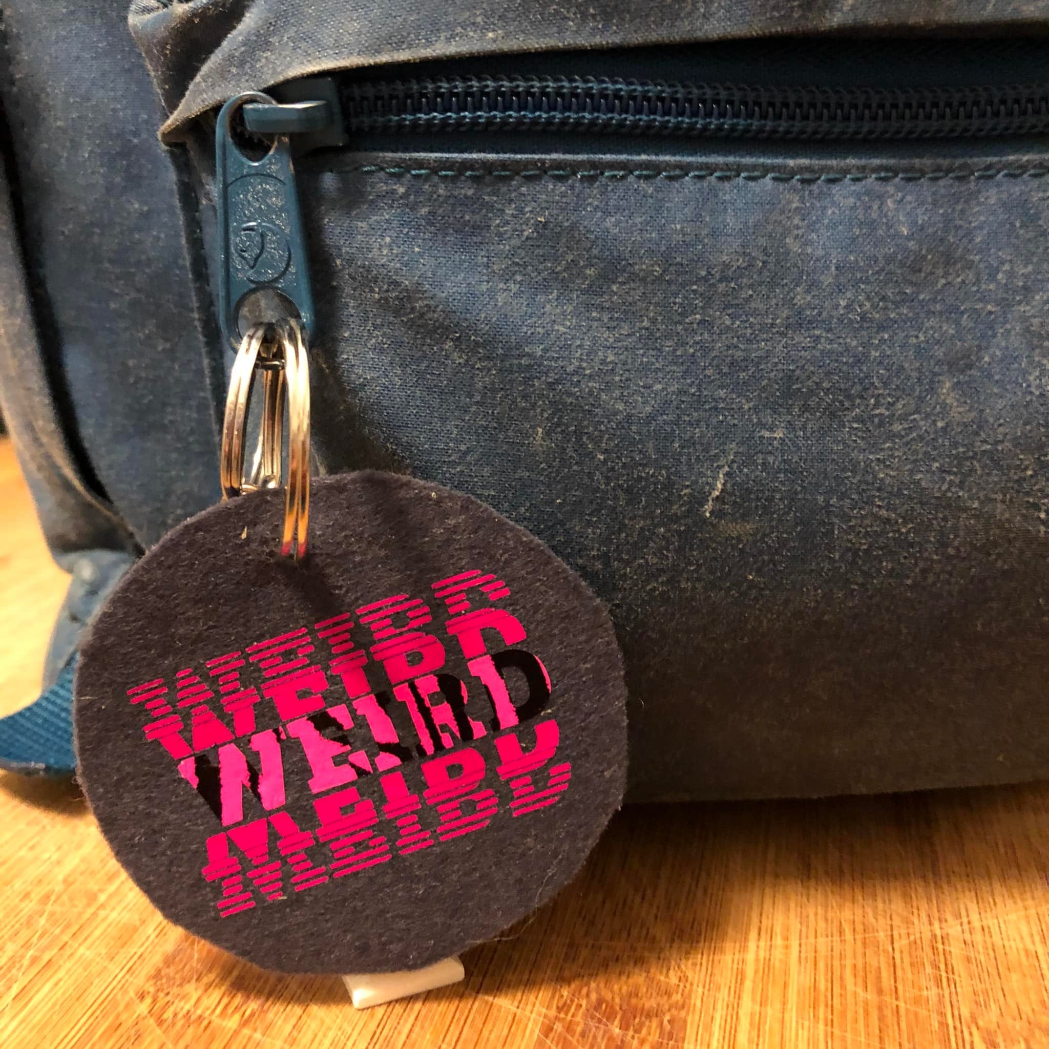 Ein Schlüsselanhänger aus Filz mit dem künstlerischen Schriftzug "WEIRD" in Pink, befestigt an einer blauen Tasche, gestaltet mit der DIY "Weird" Plotterdatei von easy-sticks.