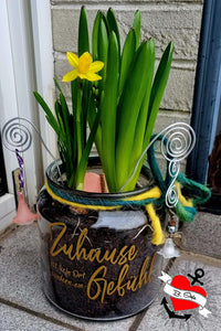 Ein dekorativer Blumentopf mit dem Aufdruck 'Zuhause ist kein Ort, sondern ein Gefühl' von easy-sticks, gefüllt mit frischen Pflanzen und bunten Details.