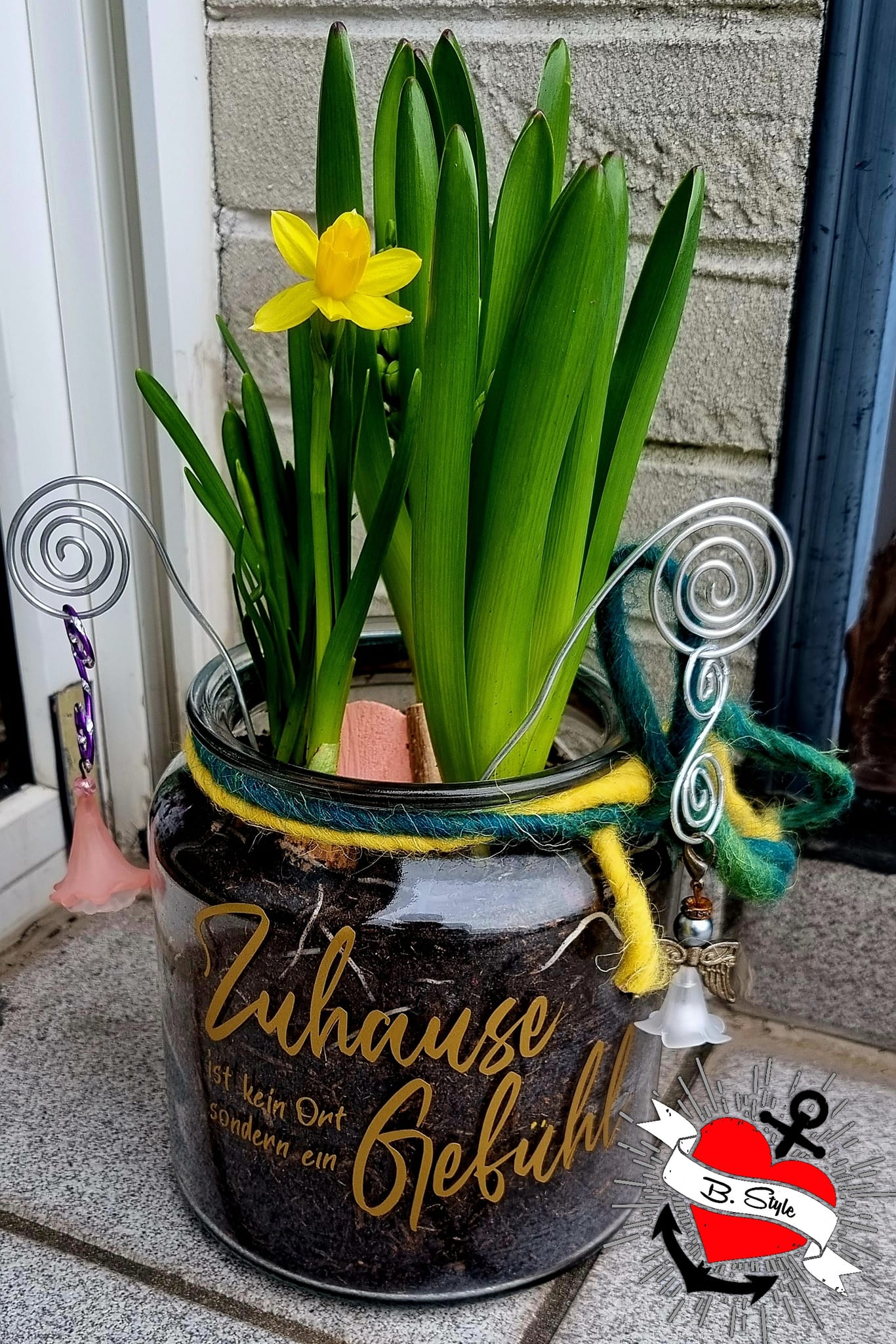 Ein dekorativer Blumentopf mit dem Aufdruck 'Zuhause ist kein Ort, sondern ein Gefühl' von easy-sticks, gefüllt mit frischen Pflanzen und bunten Details.