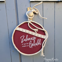 Ein stilvolles Schild mit dem Slogan 'Zuhause ist kein Ort, sondern ein Gefühl' von easy-sticks, dekorativ mit einer Schnur und an einer Holztür aufgehängt.