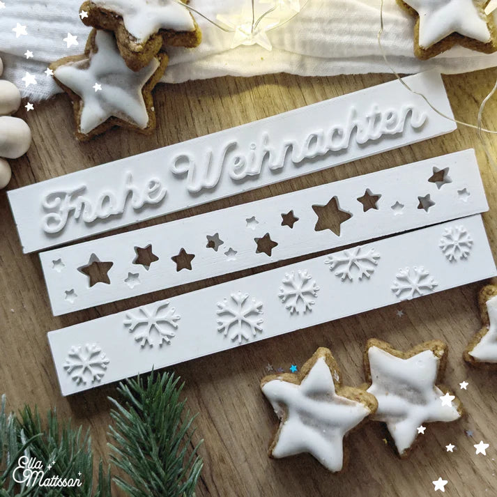 Drei Silikonformen von Easy-Sticks, darunter die Form 'Frohe Weihnachten', umgeben von Weihnachtsplätzchen und festlicher Deko.