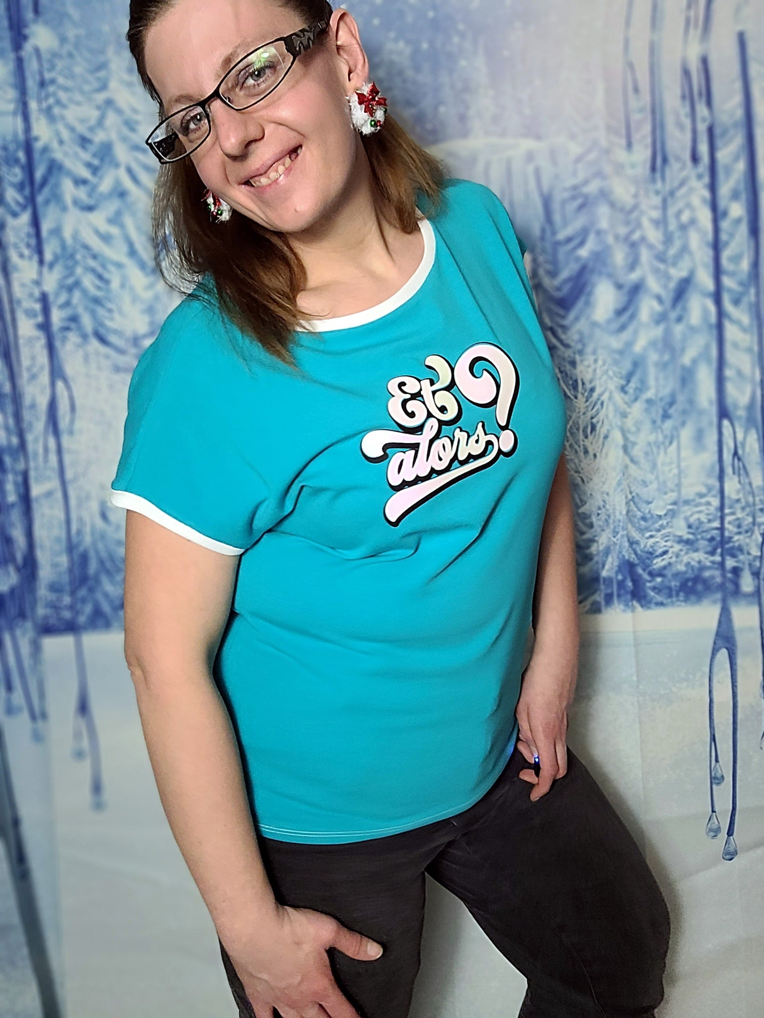 Eine lächelnde Frau trägt ein türkisfarbenes T-Shirt mit dem Aufdruck "Et alors?". Sie posiert vor einer winterlichen Hintergrundszene, die die kreative Nutzung der Plotterdatei von easy-sticks zeigt.