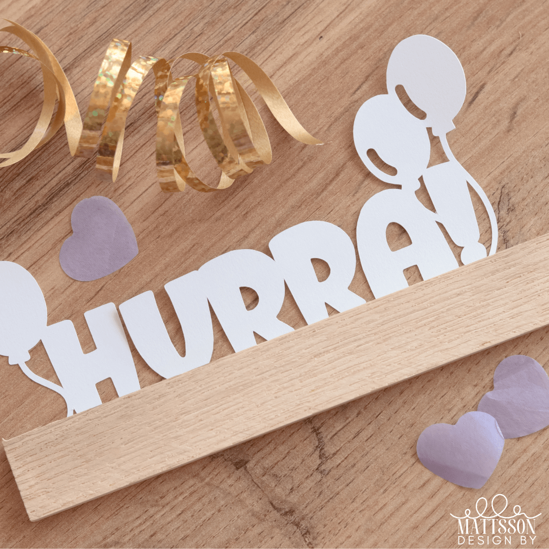 HURRA Plotter-Design aus der Kollektion von easy-sticks, präsentiert auf einem Holzuntergrund mit bunten Dekorationselementen. Ideal für kreative DIY-Projekte und personalisierte Geschenke.