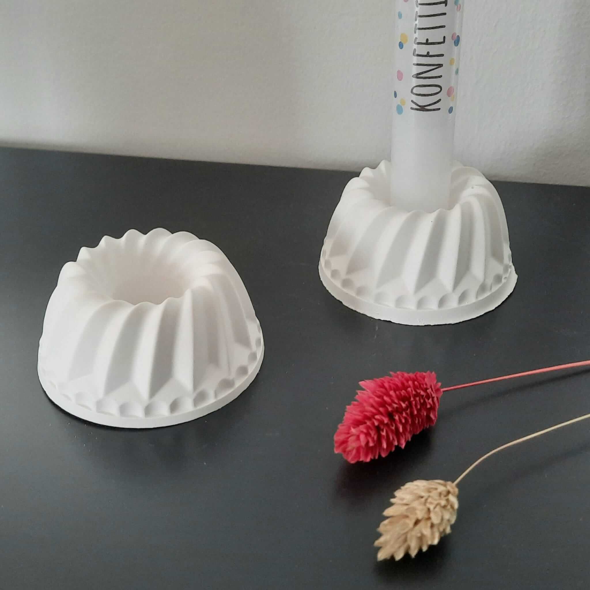 Zwei Kuchen Kerzenständer von easy-sticks auf einem Tisch, einer mit einer bunten Stabkerze, umgeben von Trockenblumen.