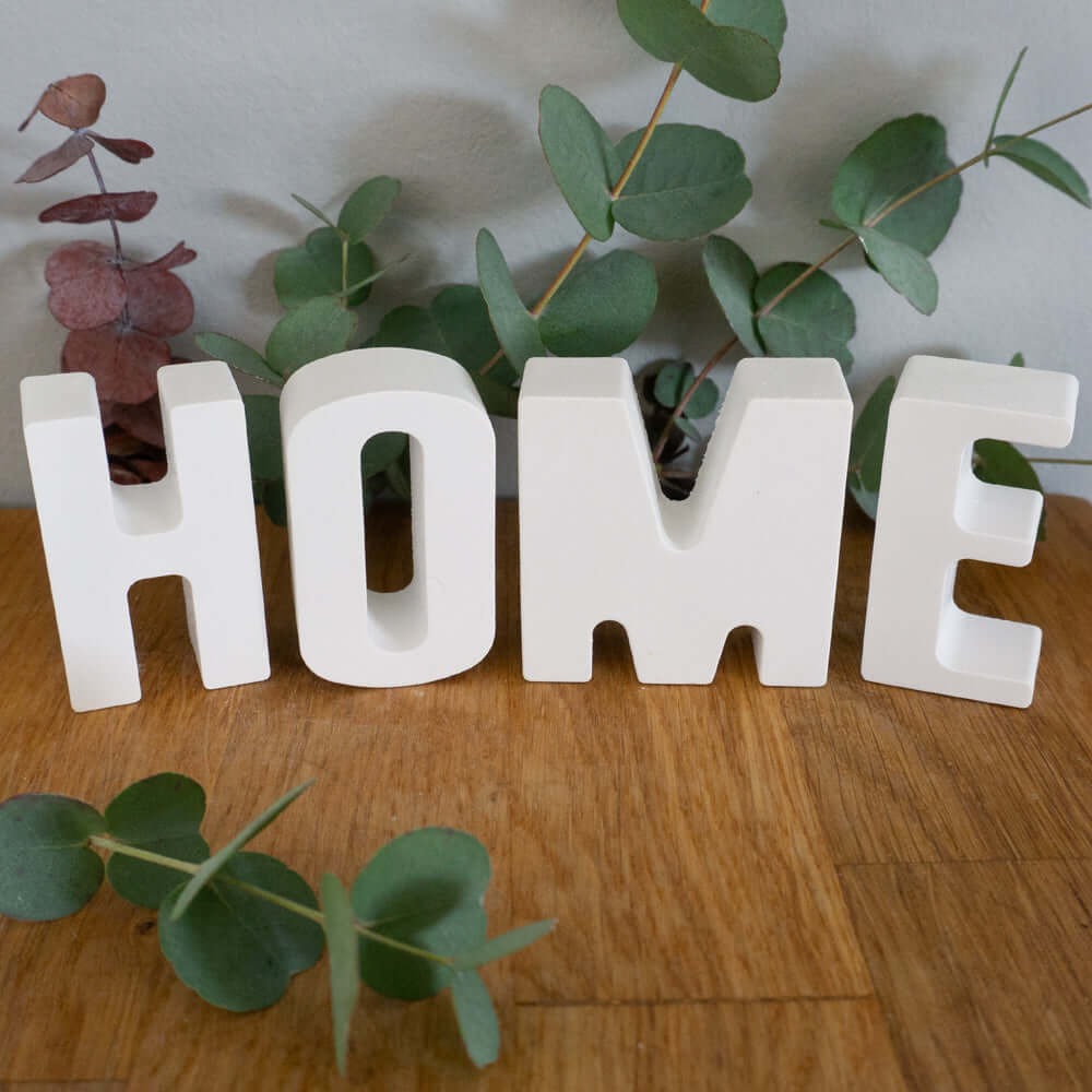 Personalisierte Buchstaben aus Kunstharz von easy-sticks, die das Wort 'HOME' darstellen, umgeben von Eukalyptuszweigen. Ideal für kreative Wohnkultur und personalisierte Geschenkideen.