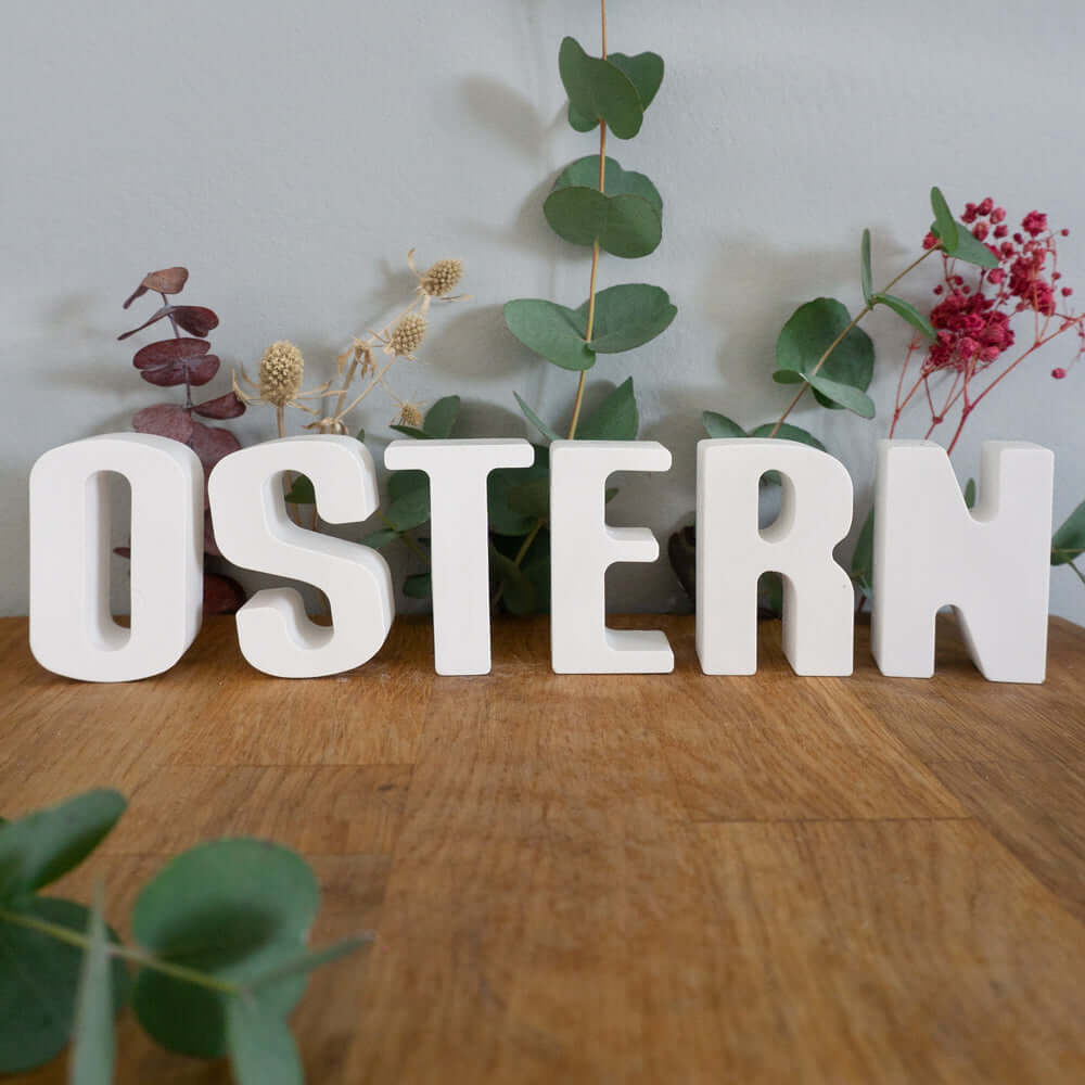 Personalisierte Buchstaben aus Kunstharz von easy-sticks, die das Wort 'OSTERN' bilden, platziert auf einer Holzoberfläche mit dekorativen Pflanzen im Hintergrund. Perfekte Dekoration für Feiertage.